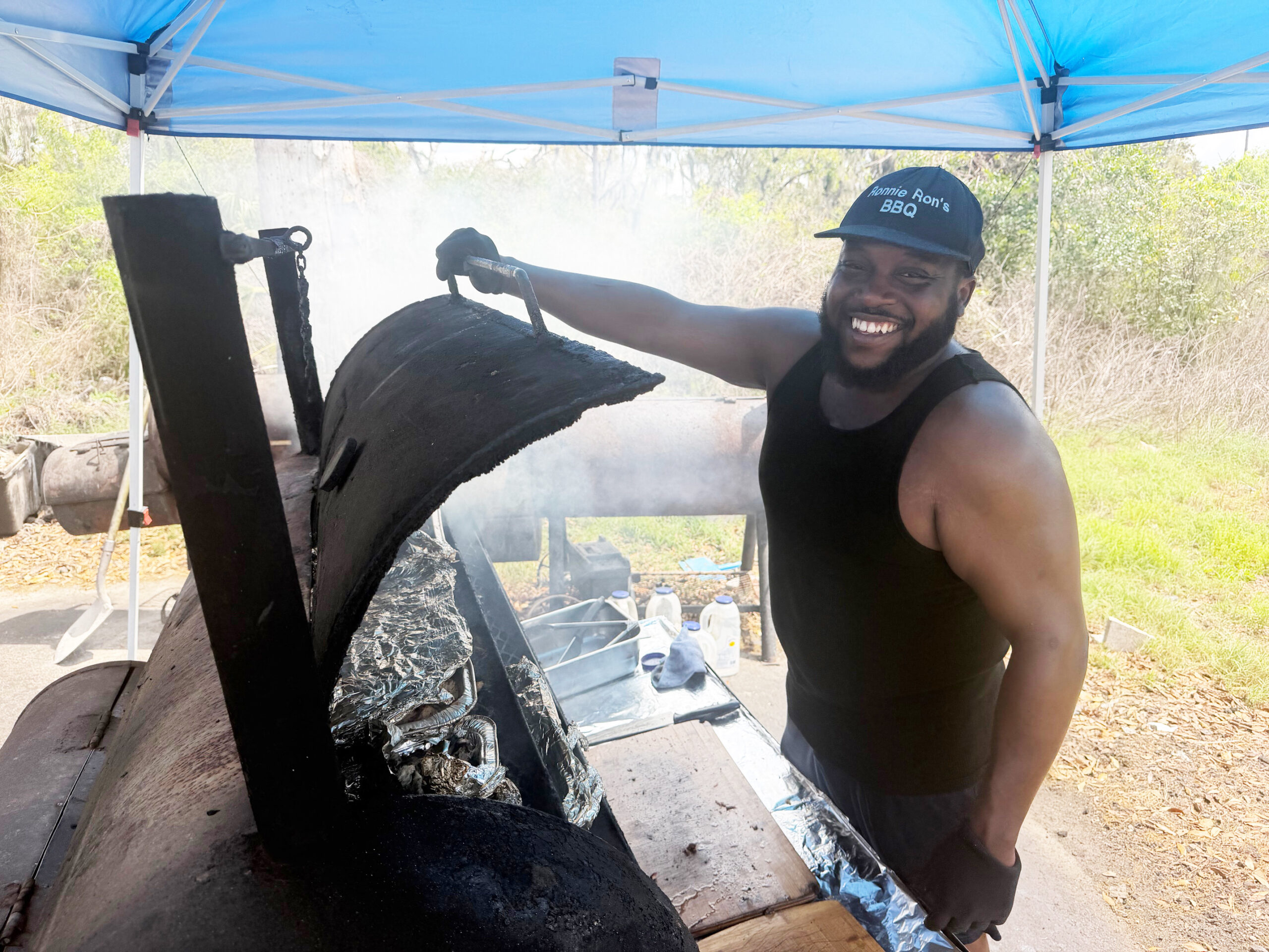 Ronnie Thomas opening his smoker with fresh smoke rolling out under a blue tent in Lithia, Florida